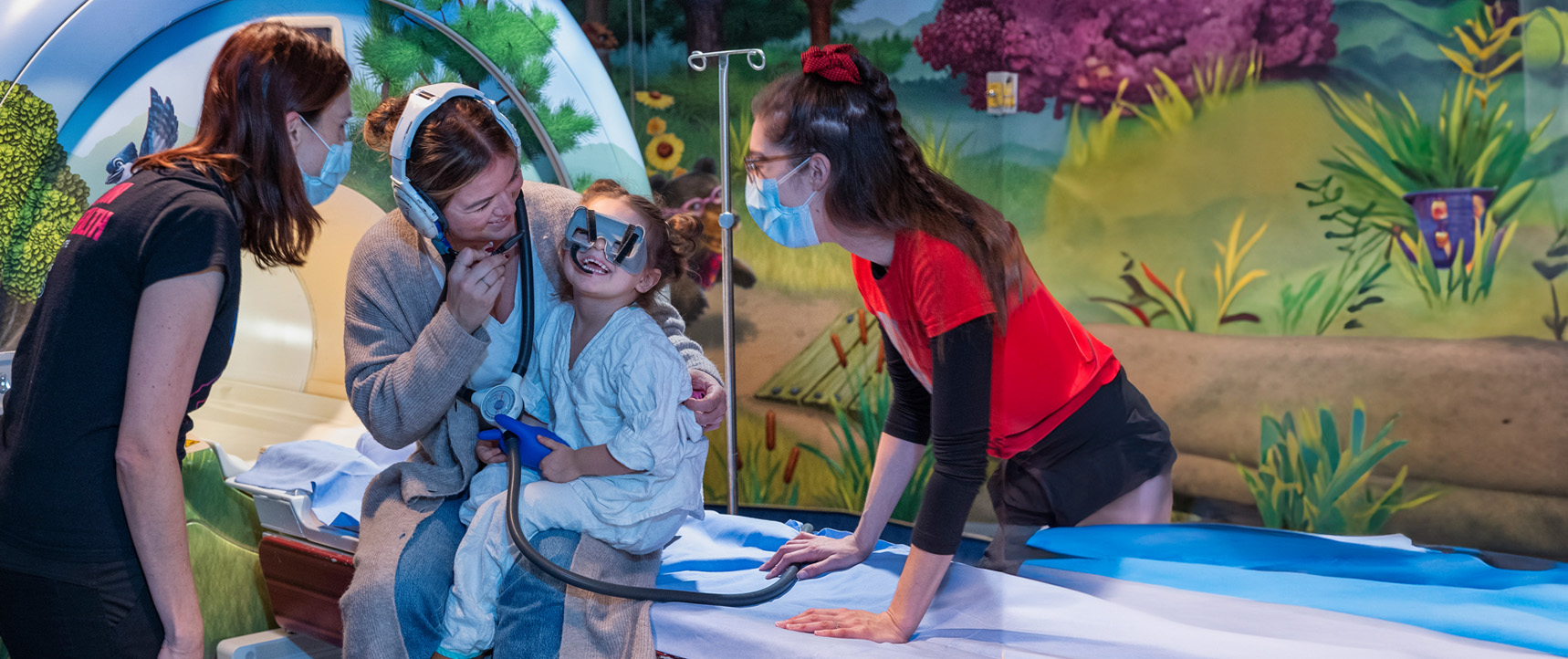 In a colourful MRI room, a child is smiling while wearing MRI simulation goggles. She is sitting on the lap of a staff member who is wearing a headset. Two other staff assist with preparing the child for the MRI.