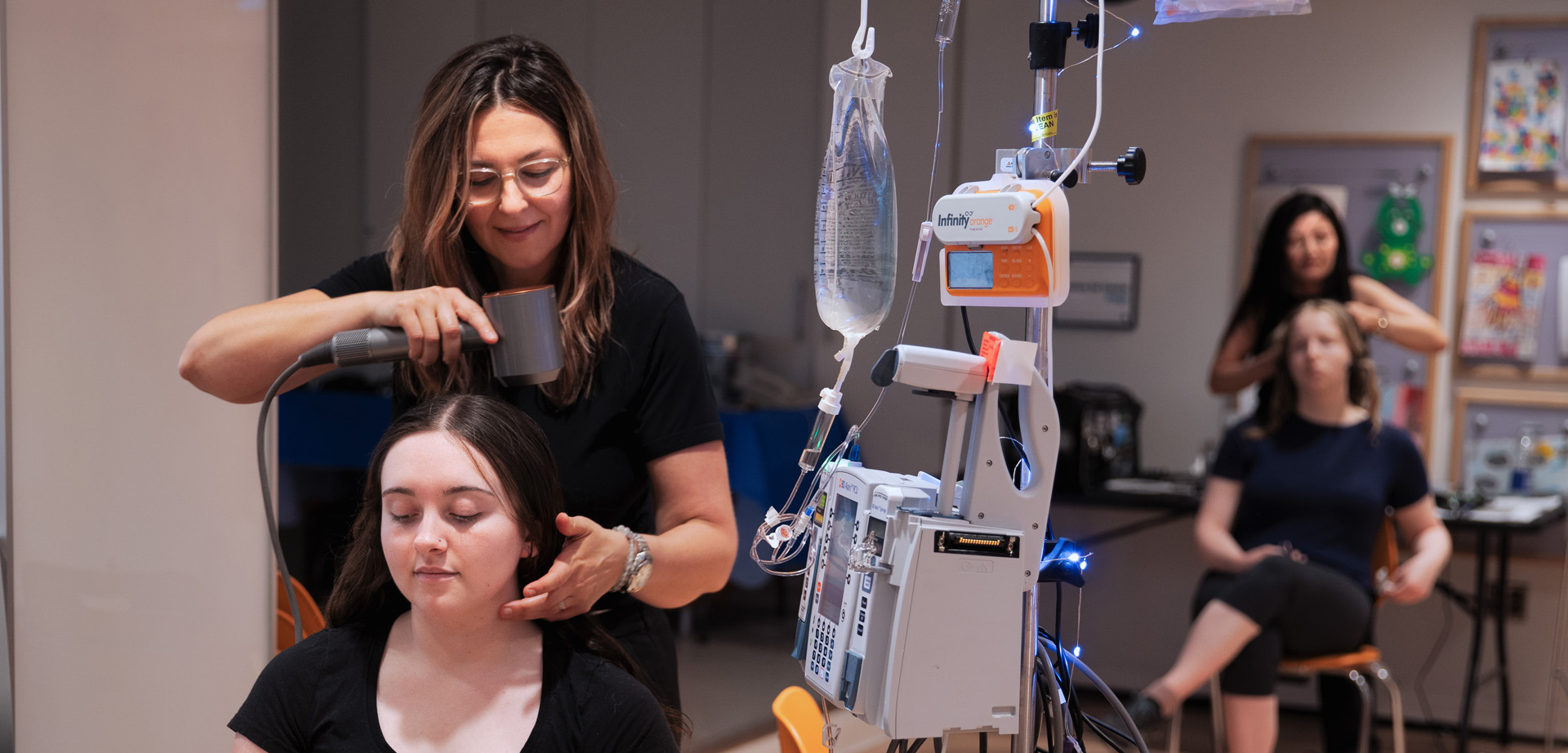 Patients getting their hair and makeup done inside Marnie's Studio. A girl is seated while connected to an IV drip. A lady is blow-drying her hair.