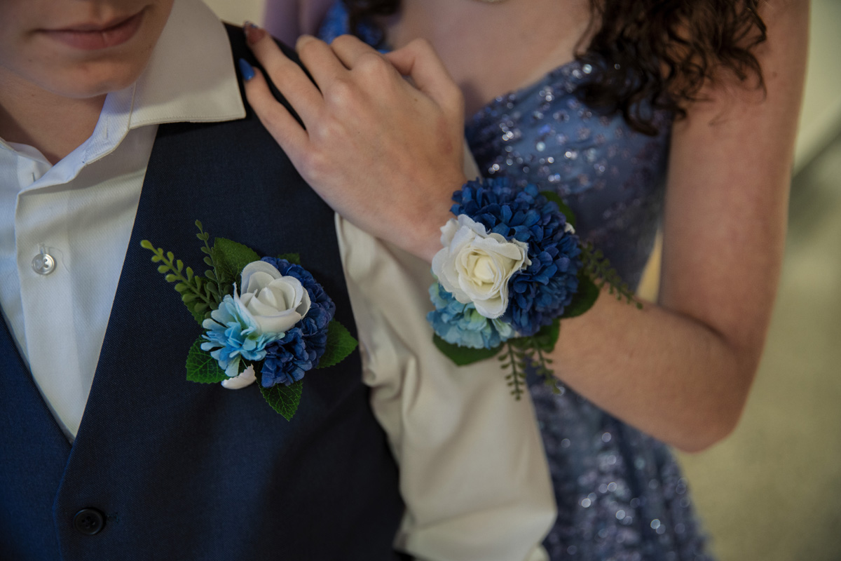 Two matching corsages consisting of white, cerulean and navy blue flowers. Rylan wears his corsage on his vest pocket, while Hope wears her corsage on her wrist.