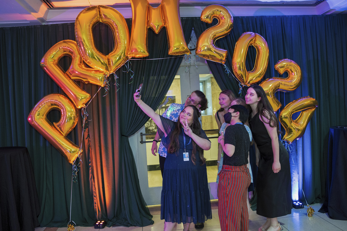 A SickKids staff member taking a group selfie with SickKids 2024 prom-goers. There are golden balloons spelling out Prom 2024 in the background.