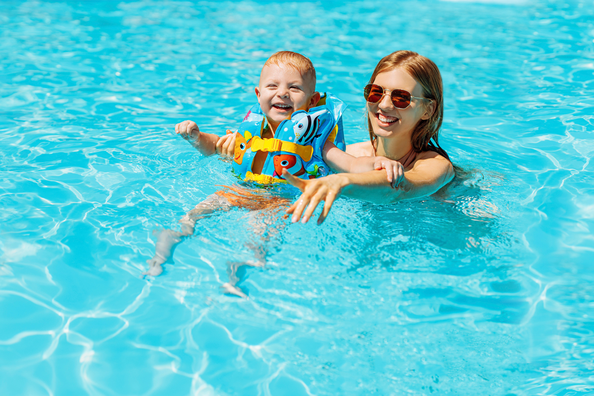 A child and mother in a swimming pool and smiling at the camera. The child is wearing a life jacket.