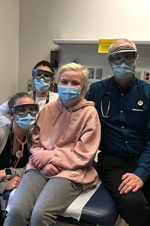 A young girl sits on a hospital bed surrounded by some of her care team.