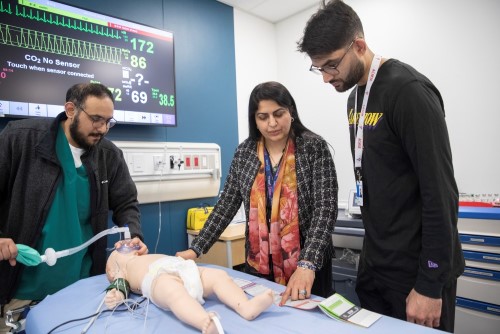Dr. Jabeen Fayyaz points to a chart to measure an infant's weight on a bed with a manikin infant wearing an oxygen mask. Two trainees watch her. A screen shows simulated vitals.