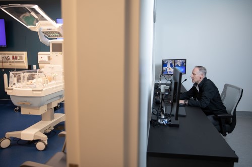 Dr. Jonathan Pirie sits in the debrief room speaking into a microphone. A wall splits the photo. On the other side of the wall is a hospital bed in the simulation room.