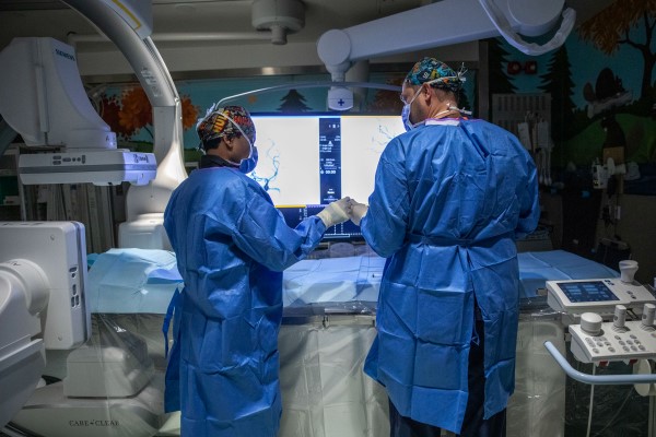 Two surgeons in blue scrubs and patterned surgical caps perform a procedure using advanced medical imaging equipment in an operating room.