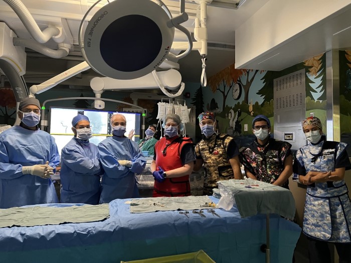 Medical team of seven professionals in surgical scrubs and masks posing in colorfully decorated operating room with overhead surgical lights.