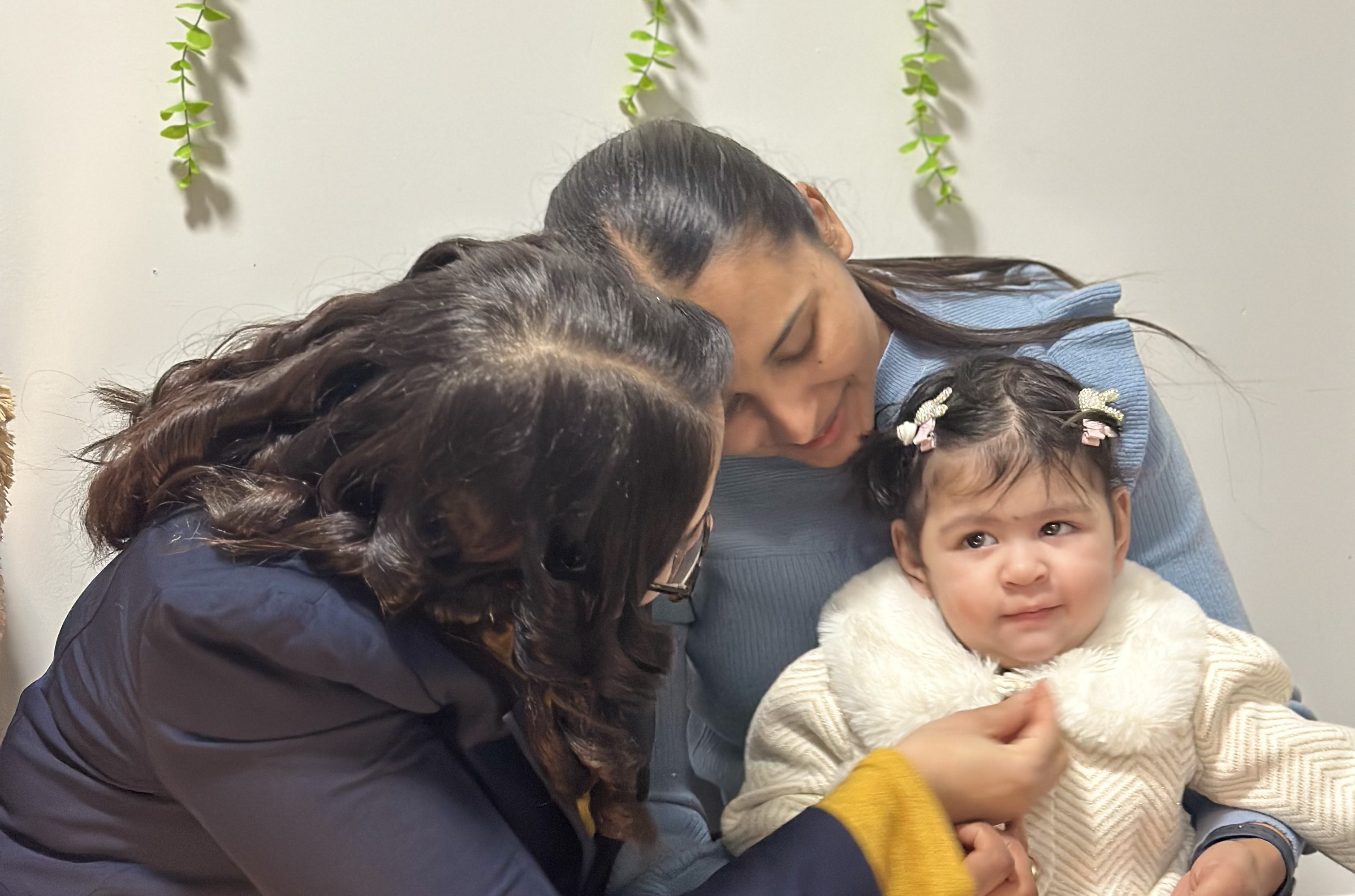 Two women holding and interacting with smiling baby in cream sweater with hair bows in comfortable home setting.