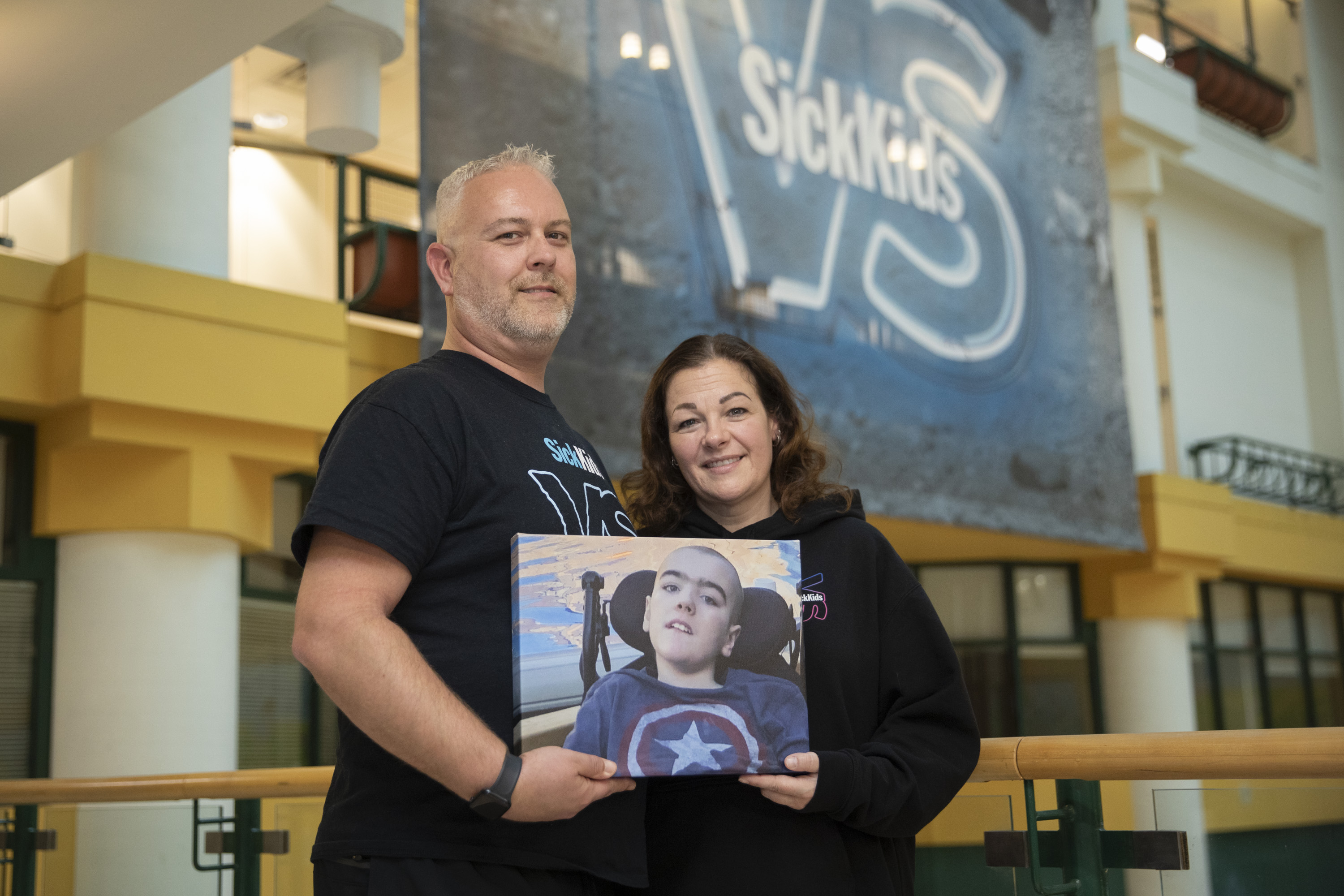 Standing in front of a blue SickKids VS banner, Doug and Melissa hold a printed photo of Noah, with the reflection of light in the pool behind him.