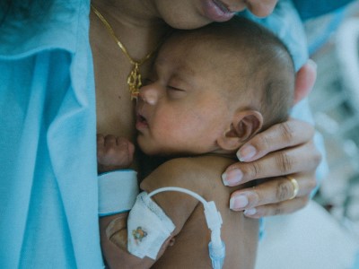 Person holding a baby with medical equipment, including arm bandage and tube, gently supporting head and back.
