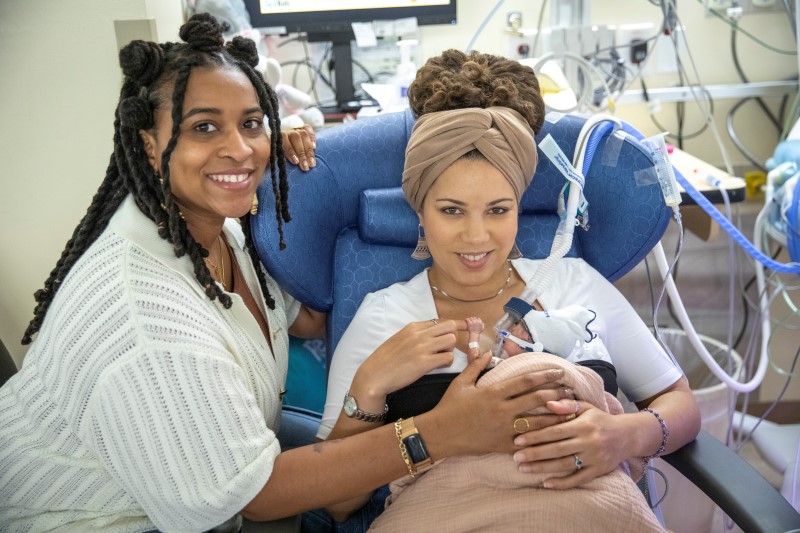 Two adults with baby in hospital; one seated holding baby with medical equipment, the other standing with supportive touch.