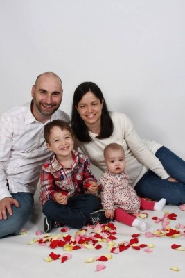 Krista, her husband, her son, and Leah, sit with a white background and red flower petals on the ground.