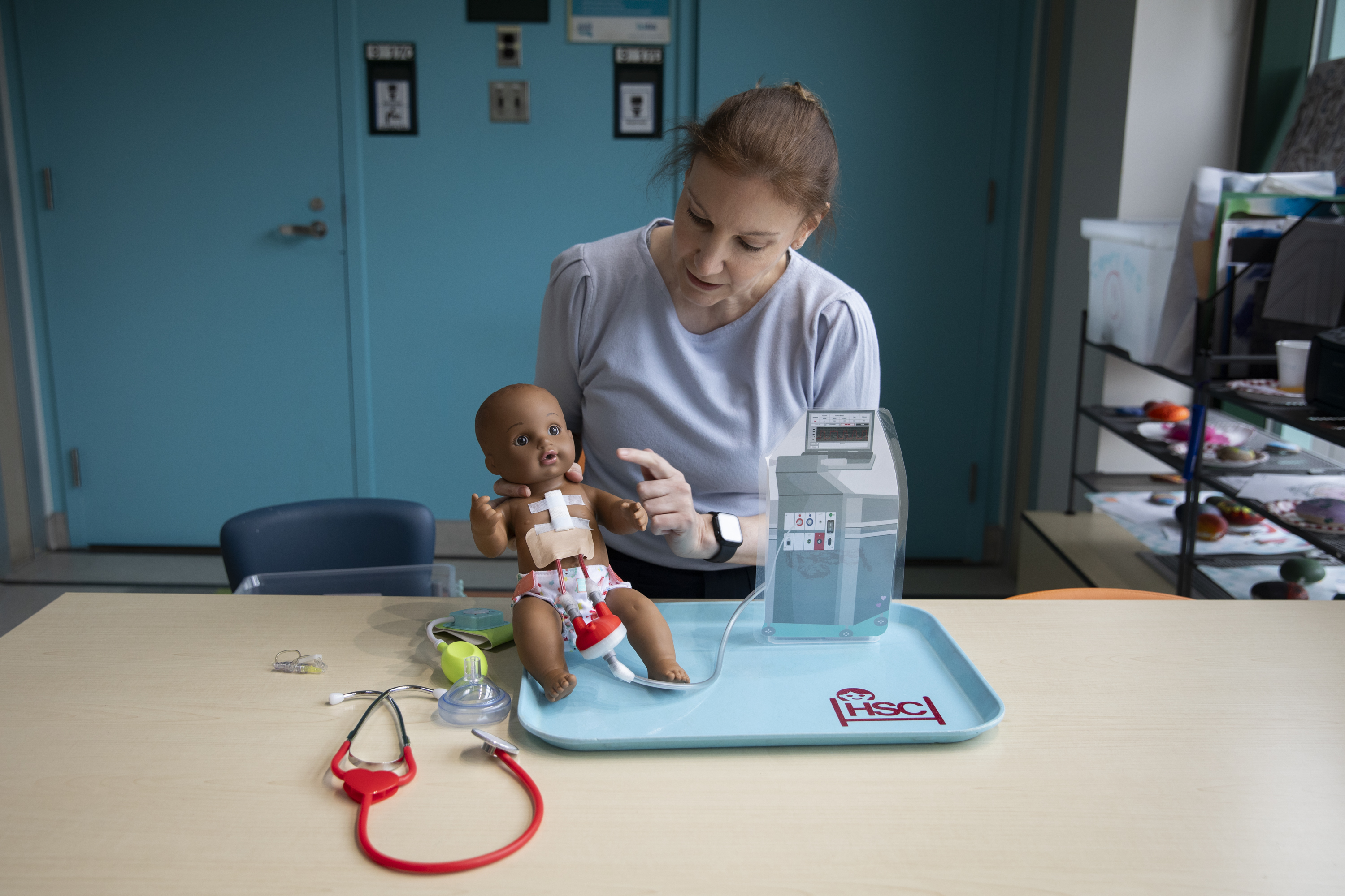 Natalie sits at a table and points at a doll that has a Ventricular Assist Device. There is a stethoscope and other medical equipment on the table.