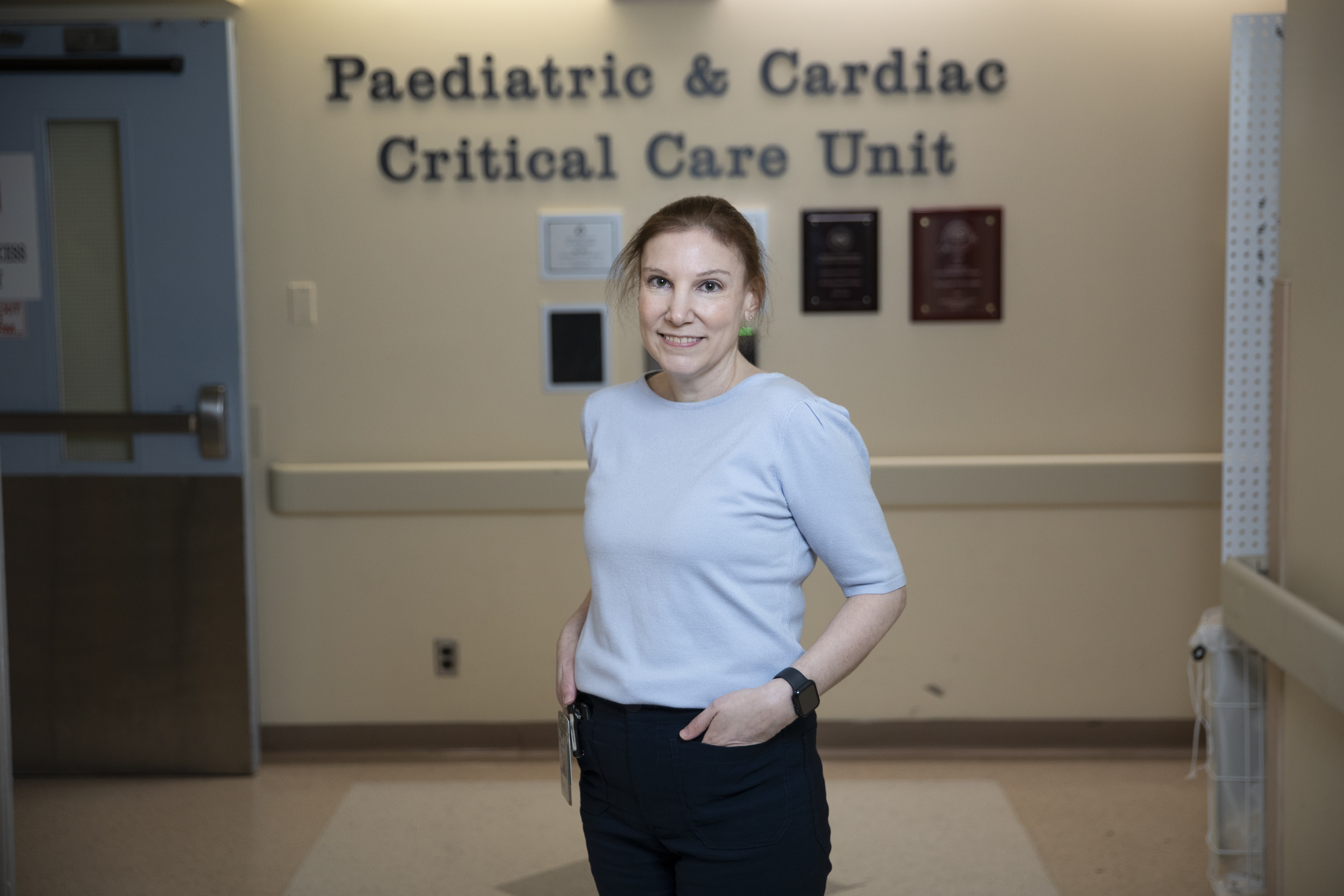 Natalie, in 2025, stands in front of the sign in the unit entrance that reads Paediatric & Cardiac Critical Care Unit.