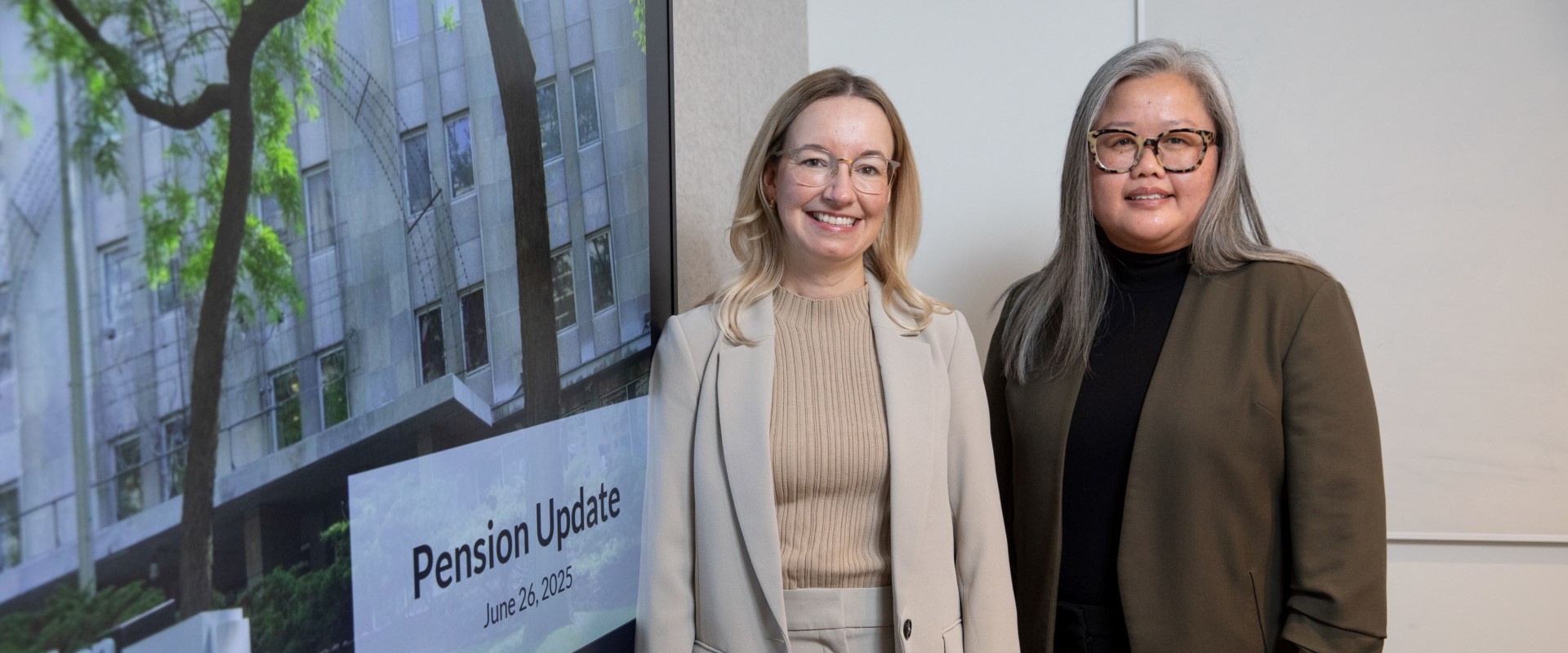 Two people standing beside a large screen displaying “Pension Update June 26, 2025” in a modern office setting.