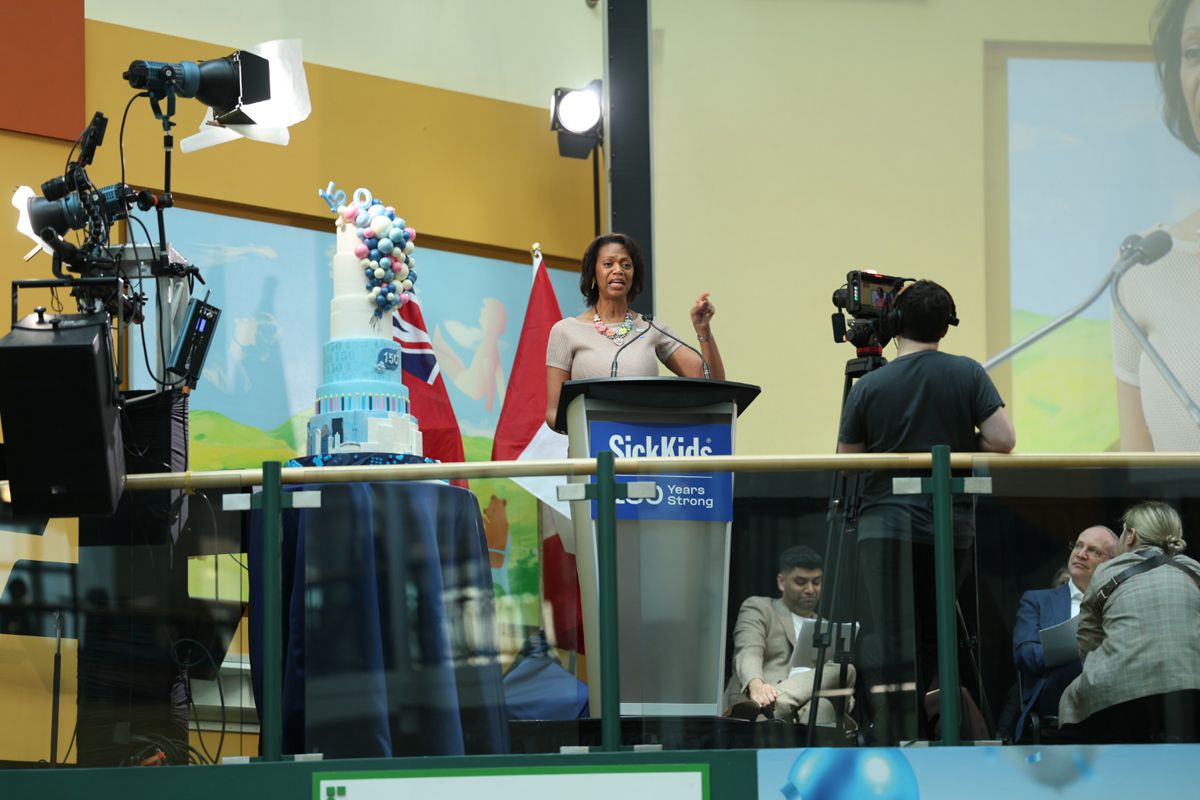Jennifer Bernard speaking at the podium on the Atrium stage next to a multilayer birthday cake.