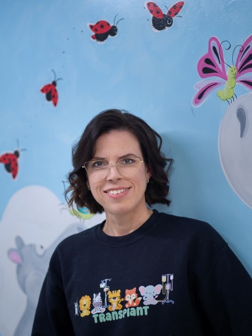 Jaina Leitch, wearing a sweater that says Transplant with cute animals on it, smiles in front of a painted mural of flying ladybugs on a blue background. 