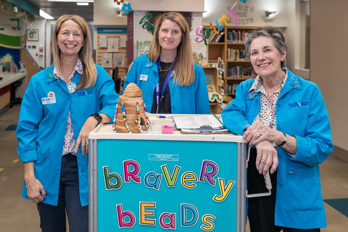 Three women in blue jackets stand behind a cart labeled 'Bravery Beads'.