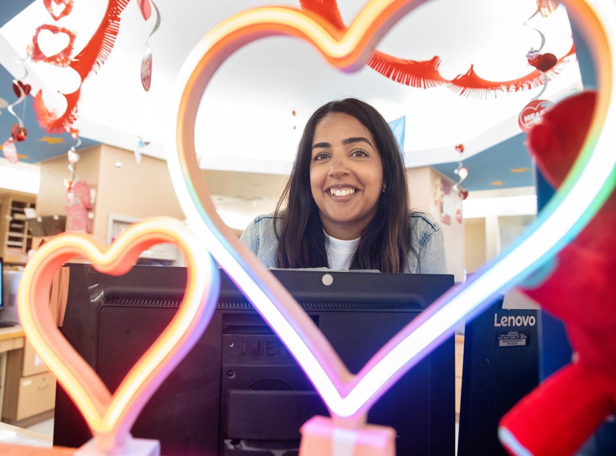 Akila stands behind the desk on 4D, her face framed by a light-up rainbow heart. 