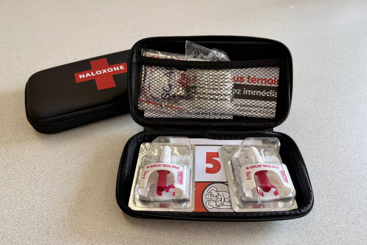 An open naloxone kit on a table shows two nasal spray doses and instructions inside a black zip case. The case is labeled naloxone.