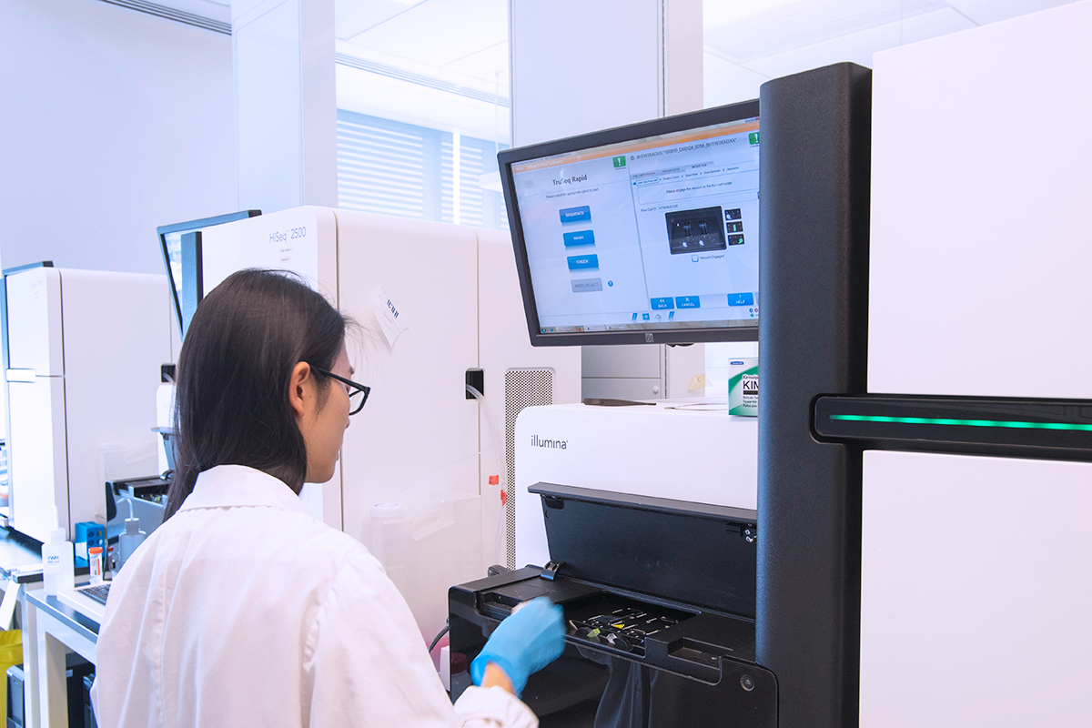 A scientist stands in a lab working at a large computer used to sequence genomes