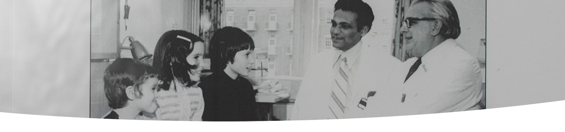 Black and white photo of Dr. Bibudhendra (Amu) Sarkar and Dr. Andrew Sass-Kortsak speaking with three children with Wilson disease