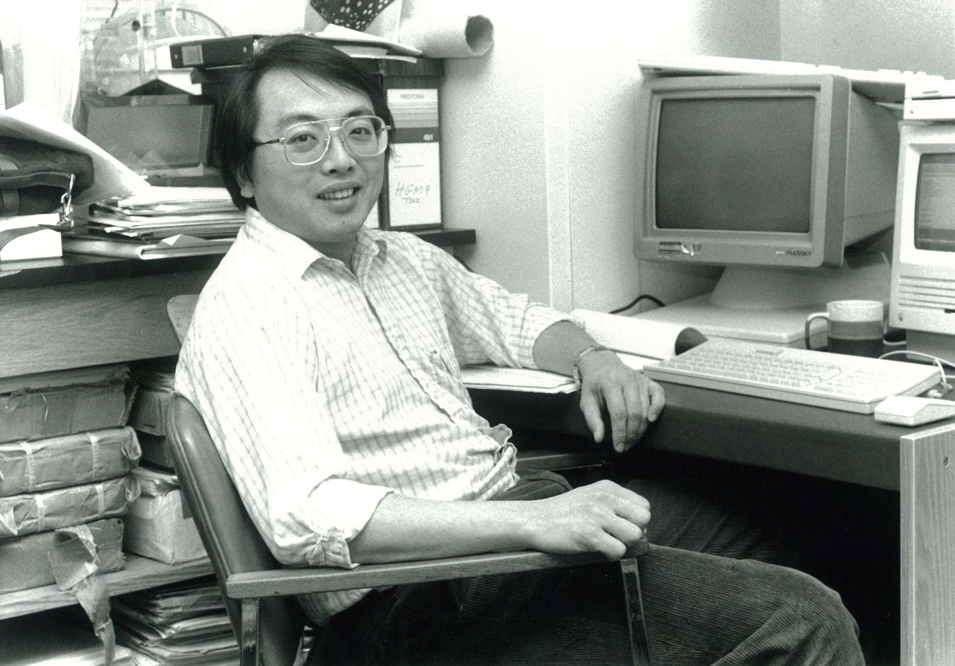 Black-and-white photo of Dr. Lapchee Tsui seated at a desk with early computer equipment, papers, and files in an office setting.