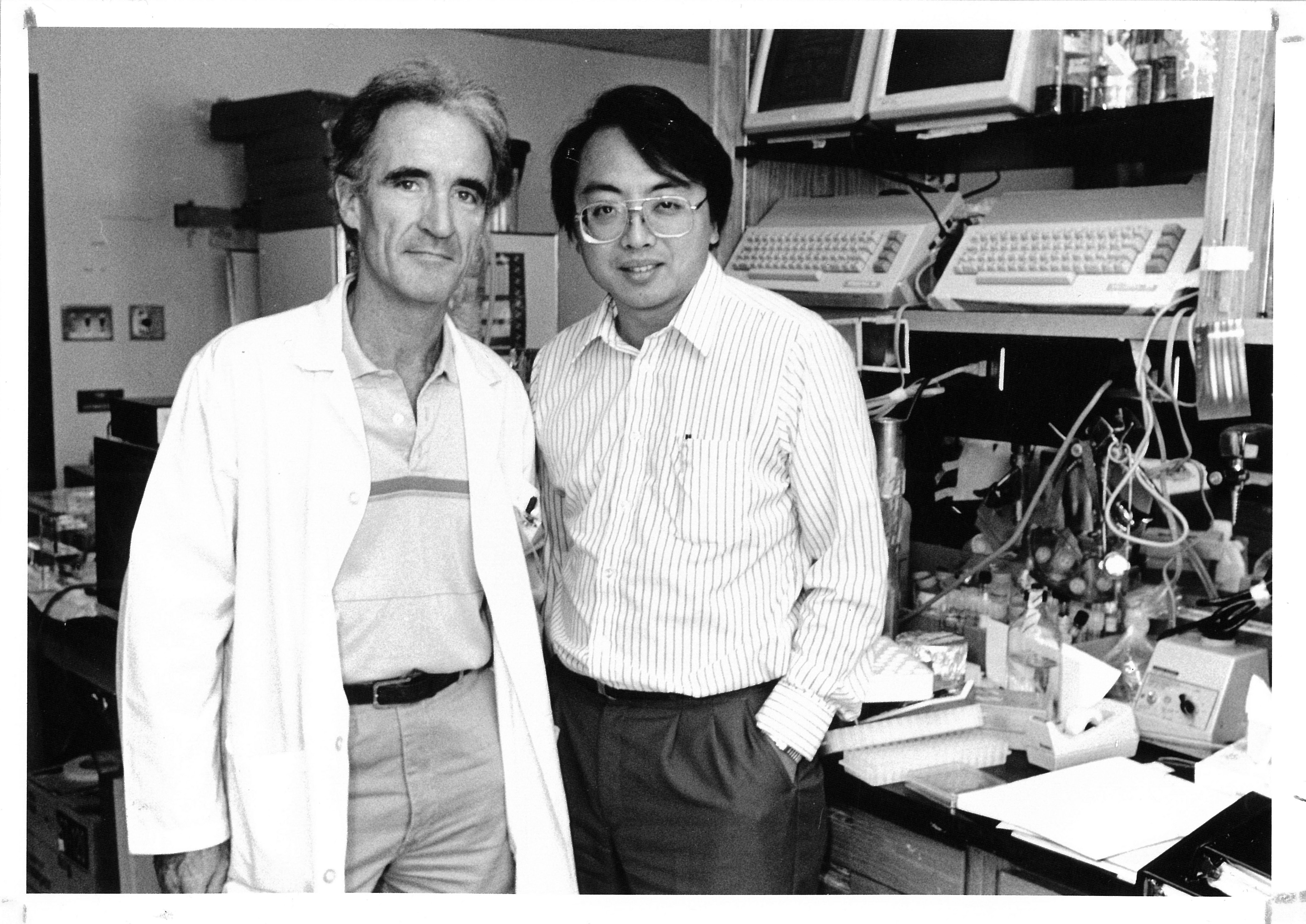Black-and-white photo of Dr. Lapchee Tsui and Dr. John Riodan standing together in a laboratory surrounded by scientific equipment and computer terminals.