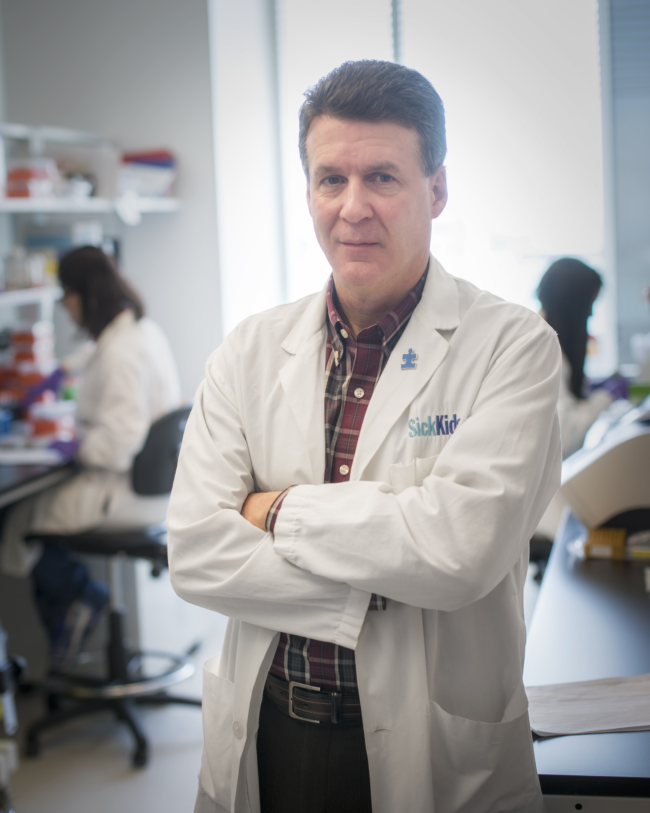 Dr. Stephen Scherer in a white SickKids lab coat leaning on a railing inside a bright, modern research building with a staircase in the background.
